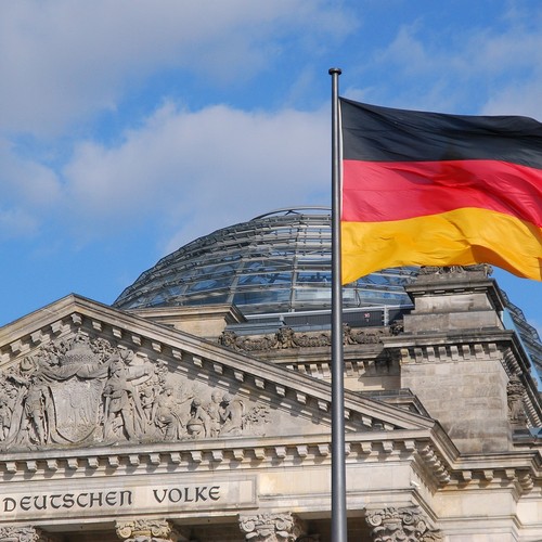 Auf dem Bild ist der Deutsche Bundestag sehen. Im Vordergrund befindet sich eine Fahne in den Farben Schwarz, Rot und Gold.