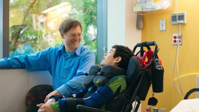 Das Bild zeigt einen jungen Mann im Rollstuhl mit einem Betreuer im Krankenhaus. Quelle:  © stock.adobe
