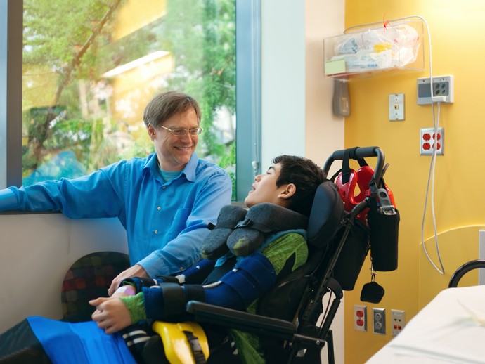 Das Bild zeigt einen jungen Mann im Rollstuhl mit einem Betreuer im Krankenhaus. Quelle:  © stock.adobe