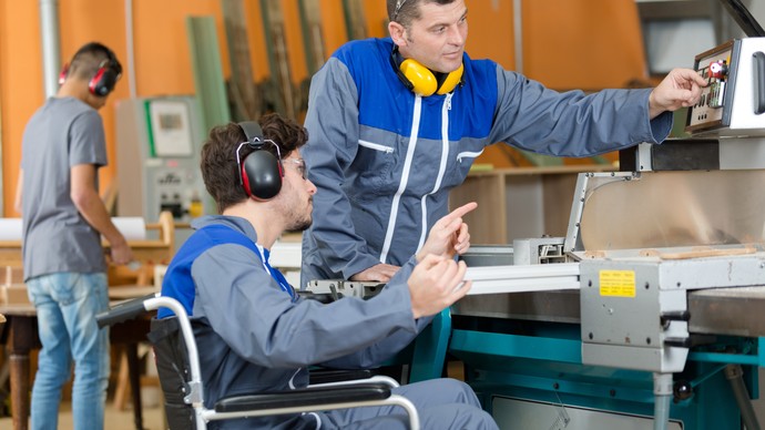 Ein Mann im Rollstuhl wird von einem anderen Mann bei der Bedienung einer Maschine eingewiesen. Quelle: © auremar/stock.adobe.com