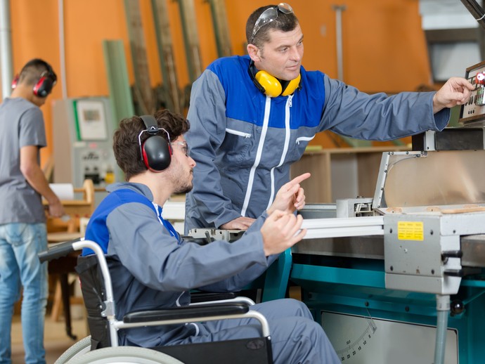 Ein Mann im Rollstuhl wird von einem anderen Mann bei der Bedienung einer Maschine eingewiesen. Quelle: © auremar/stock.adobe.com
