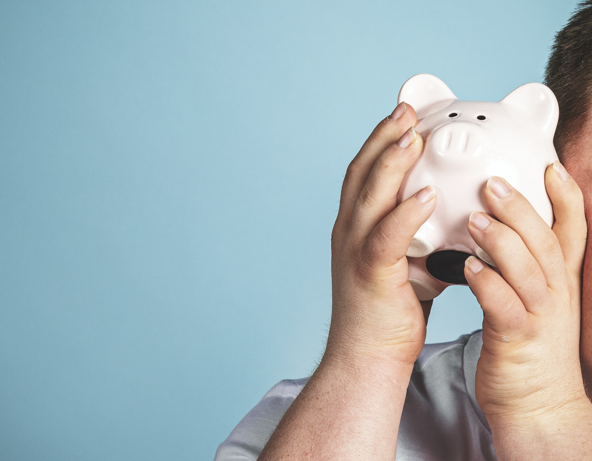 Das Bild zeigt einen jungen Mann in der Halbansicht, der ein Sparschwein neben sein Ohr hält. / Quelle: AdobeStock